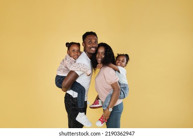 Time With Family. Overjoyed African American Family Laughing And Posing Isolated Over Yellow Studio Wall. Cheerful Father And Mom Carrying Their Daughters On Their Back.