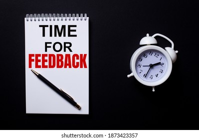 TIME FOR FEEDBACK is written in a white notepad near a white alarm clock on a black background.