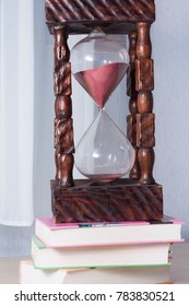 Time (for Studying) Is Running Out. A Giant Handmade Wooden Hourglass Or Sand Clock With Pink Sand On A Stack Of Three Thick Colorful School Books.