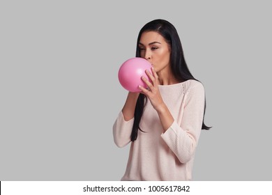 Time for fun! Studio shot of attractive young woman in casual wear blowing up a pink balloon while standing against grey background