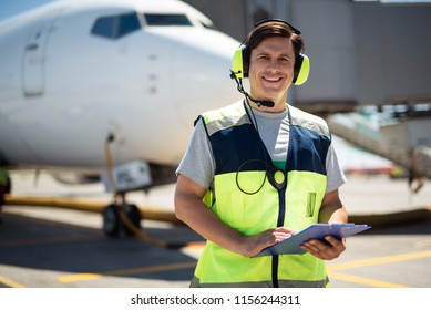 Time For Fun. Waist Up Portrait Of Smiling Worker At Airport. Man Holding Clipboard And Wearing Headphones With Microphone