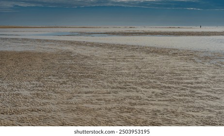 The time of low tide in the ocean. A pattern of sandy ridges formed on the endless exposed seabed. A tiny silhouette of a man walking in the distance. The blue sky. Madagascar. Morondava. 