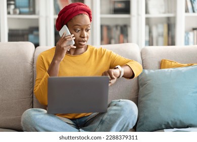 Time management concept. Worried distressed young black woman in casual entrepreneur check smart watch, sitting on couch with laptop, have phone call, working from home, getting late for meeting - Powered by Shutterstock