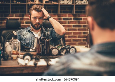 Time For New Haircut. Handsome Young Bearded Man Looking At His Reflection In The Mirror And Keeping Hand In Hair While Sitting In Chair At Barbershop