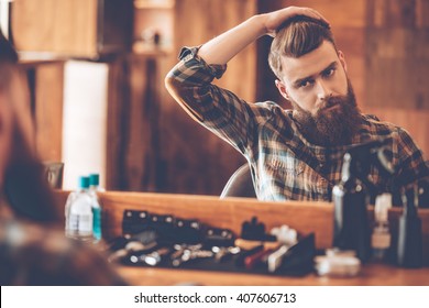 Time for new haircut. Handsome young bearded man looking at his reflection in the mirror and keeping hand in hair while sitting in chair at barbershop - Powered by Shutterstock