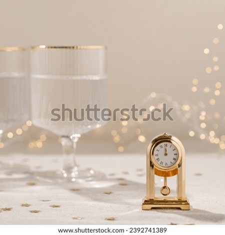 Time to New Year holiday celebration concept, gold vintage clock showing twelve o'clock, wineglasses with sparkling wine, blurred garland lights on background. Aesthetic neutral festive backdrop.