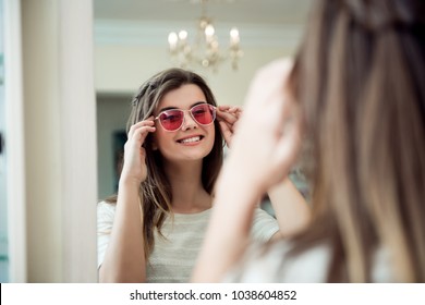It is time to refresh look with new sunglasses. Indoor portrait of attractive joyful woman on shopping, standing in optician store and trying on trendy eyewear near mirror