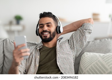 Time To Relax. Handsome Young Arab Man In Wireless Headphones Listening To Music Online On Smartphone At Home. Happy Middle Eastern Guy In Headset Watching Videos On Mobile Phone