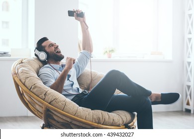 Time To Relax. Handsome Young Man In Headphones Gesturing And Keeping Eyes Closed While Sitting In Big Comfortable Chair At Home  