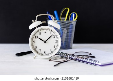 Time To Return Back To School Or Back To The Office. White Alarm Clock, Stationery. Glasses, Black Board On The Background. High Quality Photo