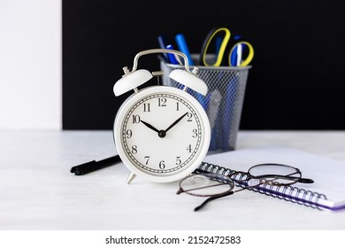 Time To Return Back To School Or Back To The Office. White Alarm Clock, Stationery. Glasses, Black Board On The Background. High Quality Photo