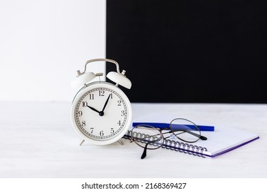 Time To Return Back To School Or Back To The Office. White Alarm Clock, Stationery. Glasses, Black Board On The Background. High Quality Photo