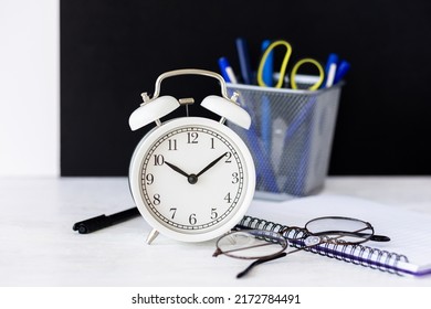 Time To Return Back To School Or Back To The Office. White Alarm Clock, Stationery. Glasses, Black Board On The Background. High Quality Photo