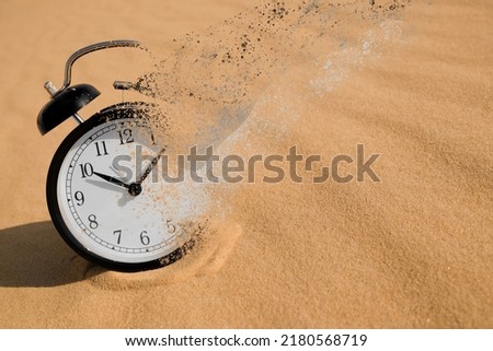 Time is running out. Black alarm clock vanishing on sand in desert