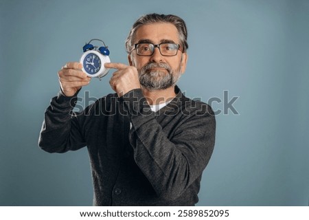 Time is running out, holding clock. Senior man is standing in the studio against background.