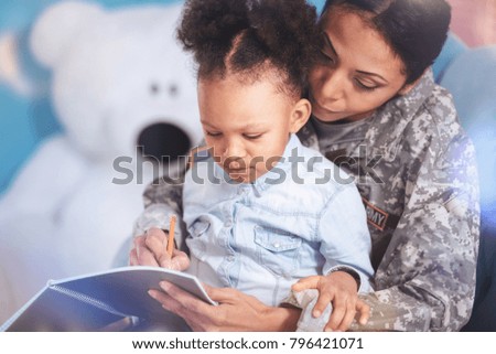 Time for studying. Positive smart nice woman holding a notebook and writing in it while studying with her daughter