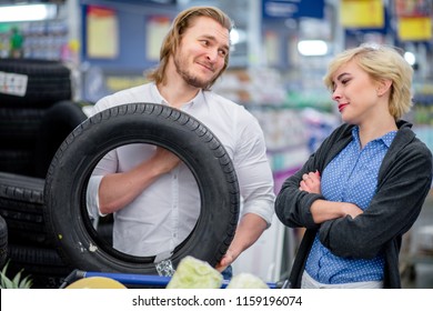 It Is Time To Swap Tires On The Car, Handsome Man With Funny, Joking Begging Expression Asks His Wife To Purchase New Tires In Big Trade Centre .