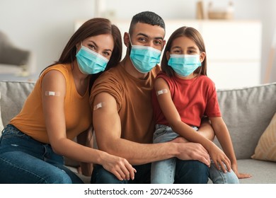 Time To Vaccinate. Happy Middle Eastern Family In Face Mask Showing Vaccinated Arm After Getting Antiviral Injection Together, Posing At Camera With Sticking Patch On Shoulder Sitting On Couch At Home