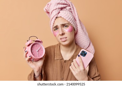 Time To Wake Up. Upset European Woman Frowns Face Doesnt Like Early Awakening Holds Alarm Clock Uses Smartphone Waits For Important Call Dressed In Nightwear Applies Hydrogel Patches Under Eyes