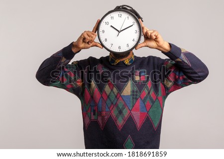 Time to work! African man hiding face behind big wall clock, afraid of deadline, wasting time, being late. Indoor studio shot isolated on gray background