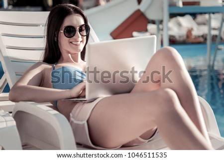 Time to work. Selective focus on a radiant millennial girl grinning broadly while relaxing on a sunbed with a laptop on her legs alone.