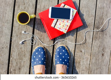 Time to work/study. Lifestyle, summer vacation, education and people concept. Young hipster girl's feet with coffee cup, smartphone, headphones, stacked books on wooden background