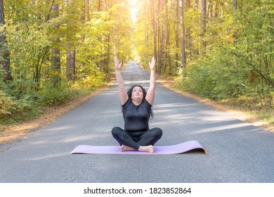 Time To Yoga! Fatty Woman Dressed In Black Tshirt And Black Sportive Pants Is Doing Yoga On Purple Mat On The Road In The Autumn Forest, Sits Stretching Her Arms Up. Sun Salutation