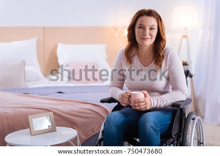 Time to yourself. Happy blond disabled woman of middle age smiling and holding a cup while sitting in the wheelchair and a bed in the background