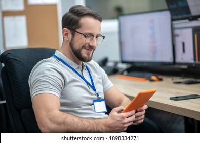 Time for yourself. Smiling interested man in gray tshirt with glasses with tablet in his hands sitting in chair at workplace