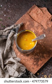 Tin Can Of Carrot Soup With Spoon, Standing On Kitchen Towel  And Clay Board Over Old Rusty Iron Background. Dark Rustic Style. Top View