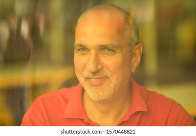 Tinted. Emotional Portrait Of A Happy And Positive Mature European Man With Bald Head Looking Down With A Smile Through A Yellow Glass At Tourists Sitting In A Summer Cafe On Vacation.Man In City