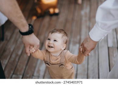 Tiny baby laughs with joy as two adults hold hands with her on wooden deck Tiny child laughs joyfully while holding hands with two adults on wooden deck in warm sunlight