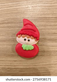 Tiny elf with red hat and green scarf sits on wooden surface. Simple design with large eyes and soft expression. Perfect for holiday-themed crafts or children's projects
