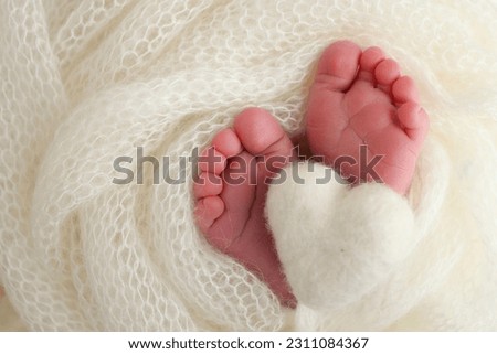 The tiny foot of a newborn baby. Soft feet of a new born in a white wool blanket. Close up of toes, heels and feet of a newborn. Knitted white heart in the legs of a baby. Macro photography 