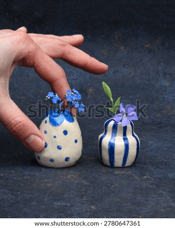 A tiny handmade ceramic bud vase with hand-painted red hearts and blue circle patterns, held between fingers to show its miniature scale. This artisanal micro vase features a rustic cream glaze and holds a small sprig of blue forget-me-not flowers. Charming handcrafted pottery for minimalist home decor, dollhouse styling, or a romantic small gift