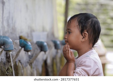 Tiny hands cup water from a rustic tap. Child drinks with quiet focus, eyes watching the stream. Wet hair clings to a soft pink shirt. Weathered concrete wall behind a row of taps