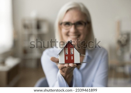 Tiny house model on hand of real estate agent woman looking at camera, smiling in blurred background. Senior realtor offering property buying, rent of apartment, mortgage consultation