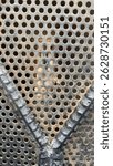 A tiny jumping spider rests on a rusted and dusty perforated metal grid with circular holes, blending naturally into the industrial texture of the surrounding environment.