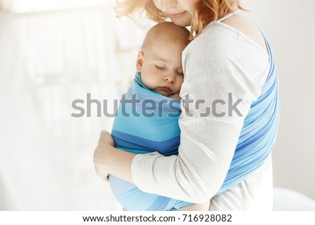 Tiny newborn child close eyes and having good sleep in baby sling feeling protection from his beautiful young mother. Family, lifestyle concept.
