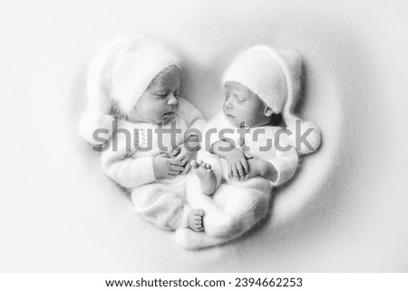 Tiny newborn twin boys in white bodysuits in white caps. Newborn twins sleep next to their brother on the background of the heart. Two newborn twin boys hugging each other. Black and white.