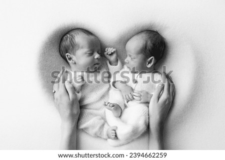 Tiny newborn twin boys in white bodysuits on a white background. Newborn twins sleep next to their brother on the background of the heart. Two newborn twin boys hugging each other. Black and white 