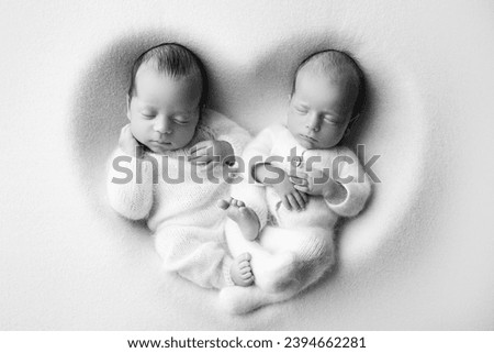Tiny newborn twin boys in white bodysuits on a white background. Newborn twins sleep next to their brother on the background of the heart. Two newborn twin boys hugging each other. Black and white. 