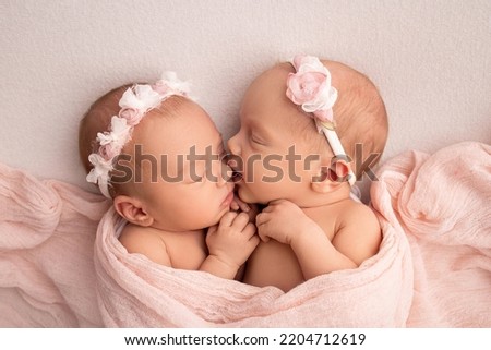 Tiny newborn twin girls. A newborn twin sleeps next to his sister. Newborn twin girls on the background of a pink blanket with pink bandages. The girls gently hug and kiss their sister in a cute pose.