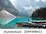 Tiny person standing on dock with colourful canoes at turquoise blue lake surrounded by mountains