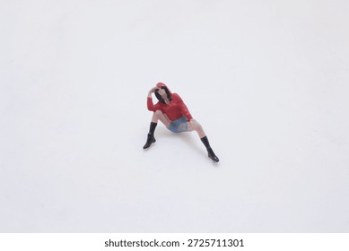 A tiny plastic figurine sits on a clean white surface, wearing a red hoodie, b