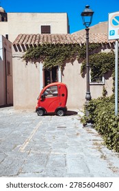 A tiny red bubble microcar parked beside a vine-covered stone house with terracotta roof on a sunlit cobbled lane in a Sardinian old town.