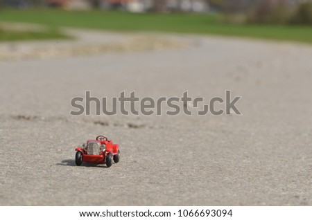 Tiny red oldtimer toy car driving down a concrete road with meadows in the background