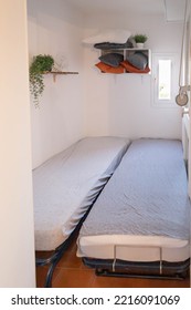 Tiny Room In A Vacation Apartment With Two Beds With Grey Sheets. Minimal Decoration. A Shelving With Some Cushion And One Plant