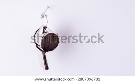 Tiny, round metal whistle hangs from a hook. Its dark surface has a textured finish. The plain white background makes it stand out. This miniature object is lightweight and portable