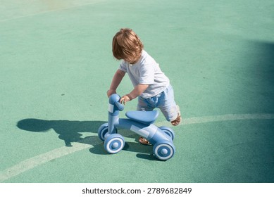 A tiny toddler boy concentrations on mounting a blue four-wheeled balance bike on an outdoor sports court. The little child in a white t-shirt and light shorts steps over the seat, casting a distinct dark shadow on the green rubber flooring next to a painted tactical marking line.